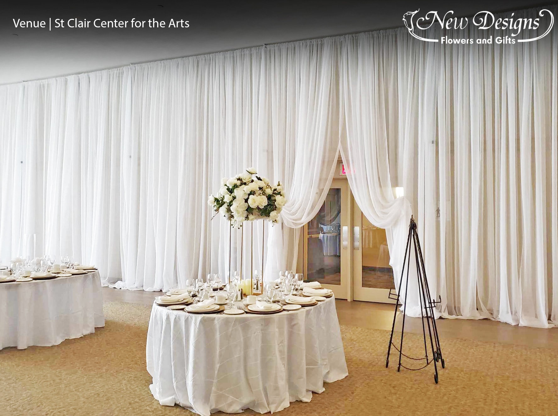 A hall overflowing with draped White Chiffon along the walls and on the tables, Elegantly pulled back to reveal an entry door. With a Tall 4 posted gold stand adorned on top with a full compact arrangement of white roses hydrangeas and orchids